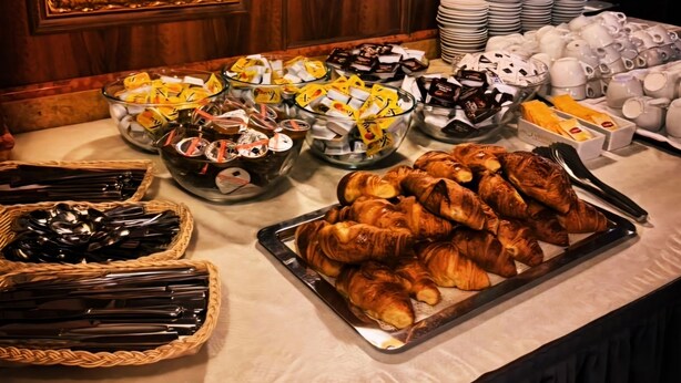 Een buffettafel met diverse gerechten en bestek, waaronder croissants en kommen met eten, op de achtergrond houten lambrisering en lijstwerken.