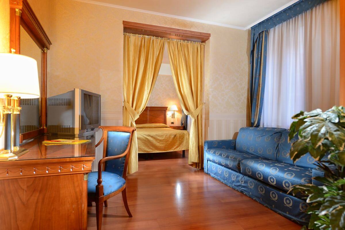 Habitación de hotel con cama, sofá azul, escritorio de madera, televisor y una planta grande.