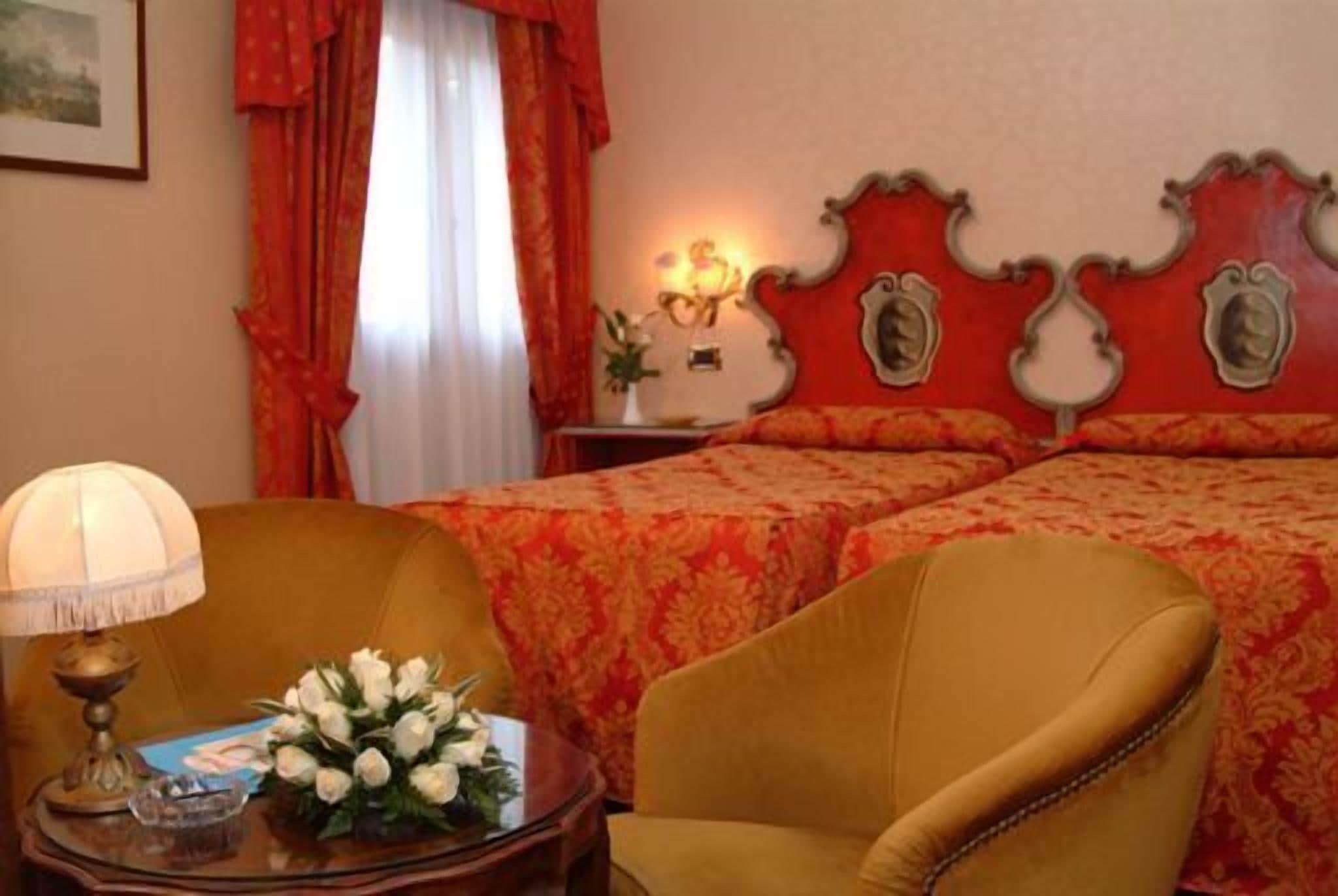 A hotel room with two beds, a sitting area with a chair, a table with a lamp, and a vase of flowers.