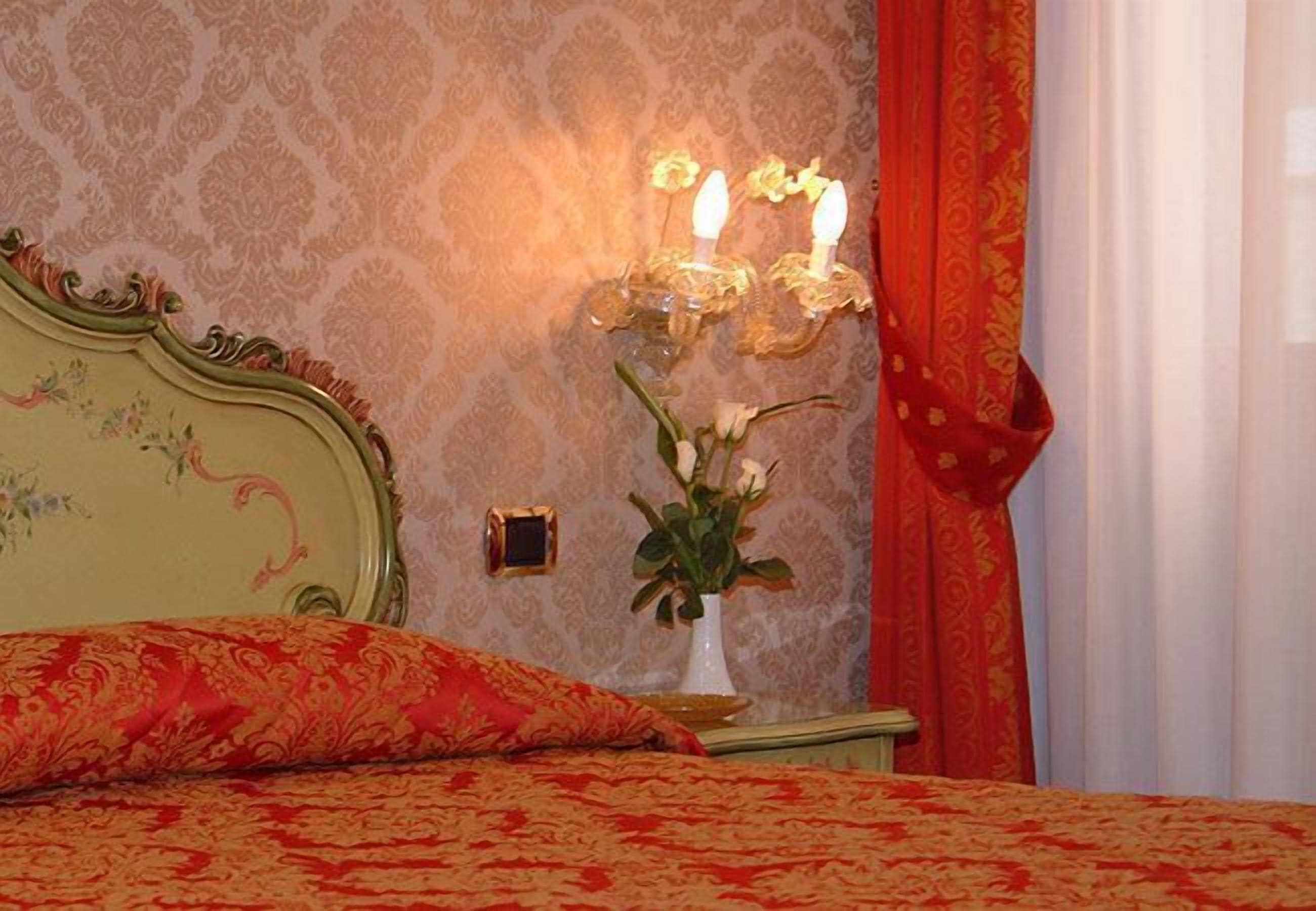 A bedroom with a floral-patterned bedspread, a decorative wall sconce, and a vase with flowers on a nightstand.