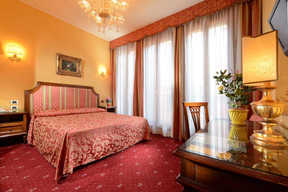 Habitación de hotel con una cama grande, un candelabro, un escritorio con lámpara y una ventana con cortinas.