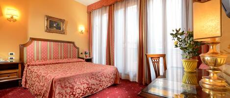 A hotel room with a large bed, a chandelier, a desk with a lamp, and a window with curtains.