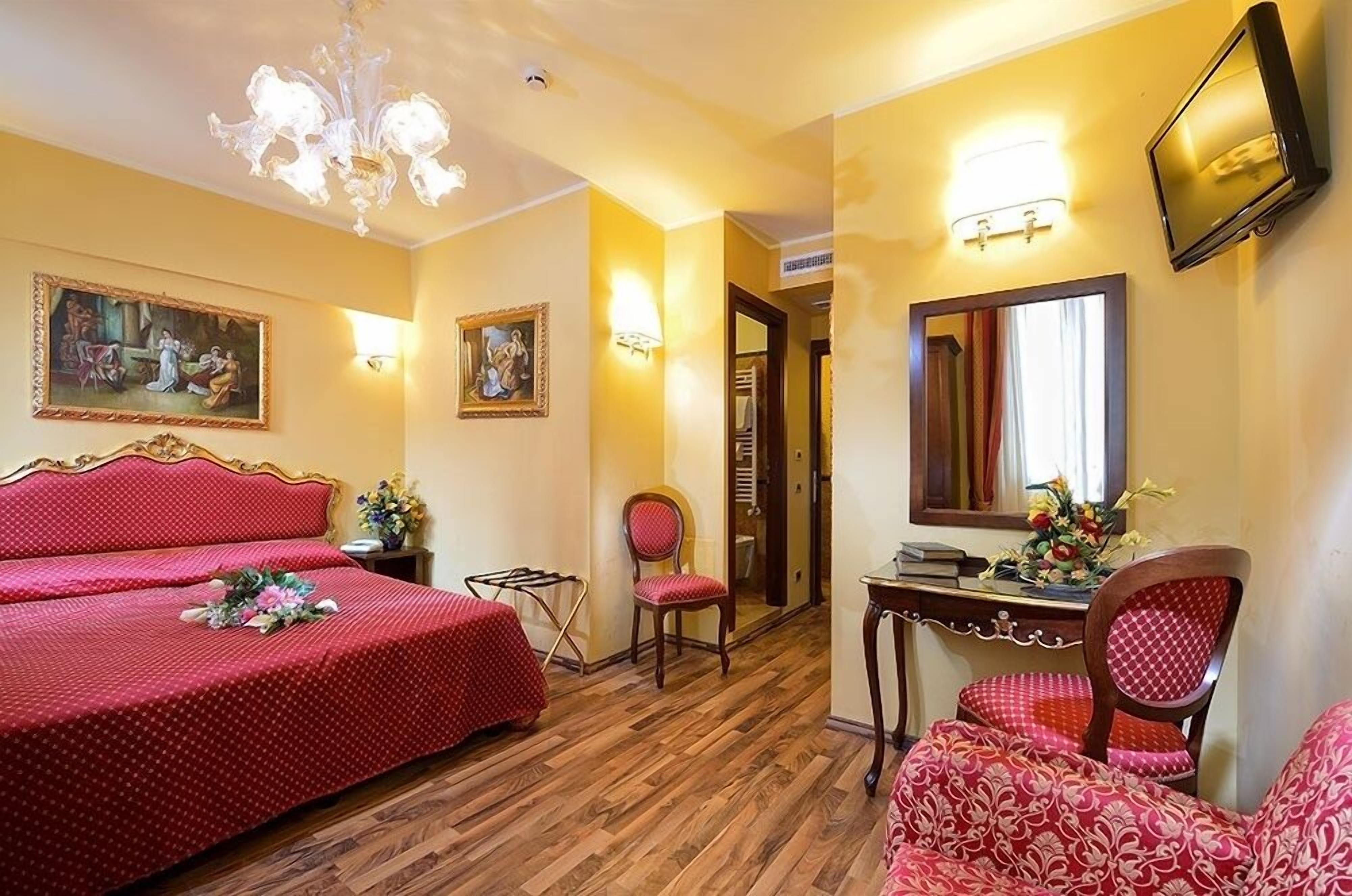 A hotel room with two beds, a desk, a chair, a TV, and artwork on the walls.