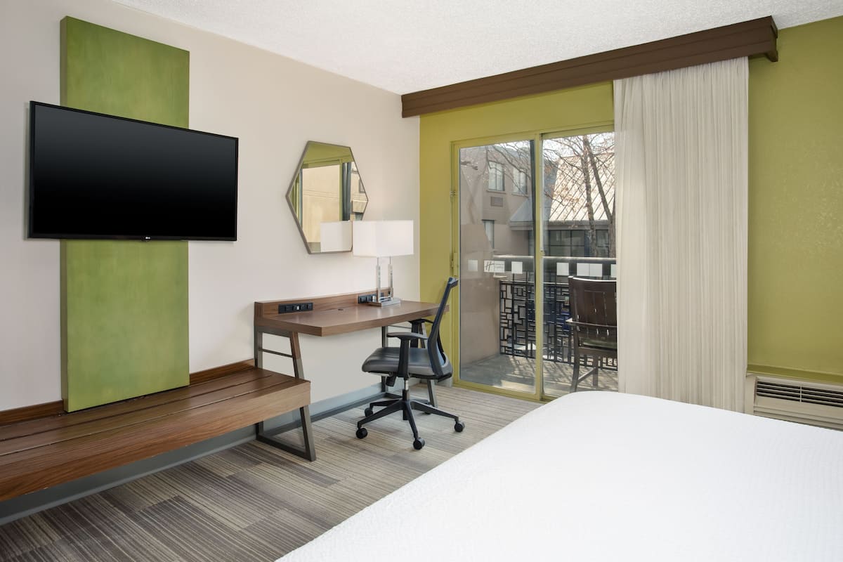 Une chambre d’hôtel comprenant un lit, un bureau avec une chaise, un téléviseur à écran plat, un miroir et une porte-fenêtre coulissante donnant sur un balcon.