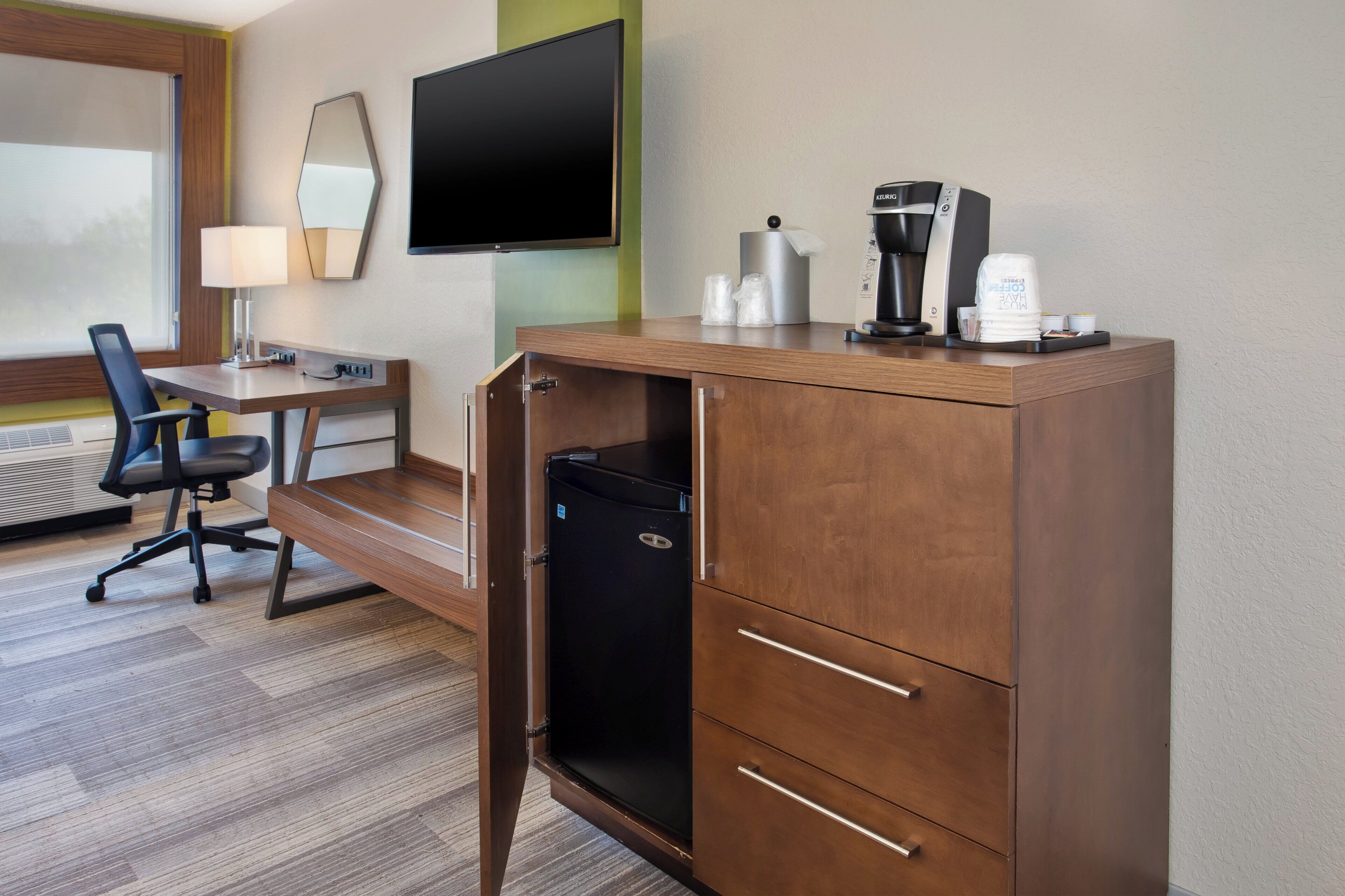 A hotel room with a wooden desk, a black chair, a flat-screen TV, a coffee maker, and a minibar.