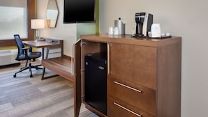 Une chambre d’hôtel avec un bureau en bois, un réfrigérateur noir, un téléviseur à écran plat, une machine à café et une chaise.