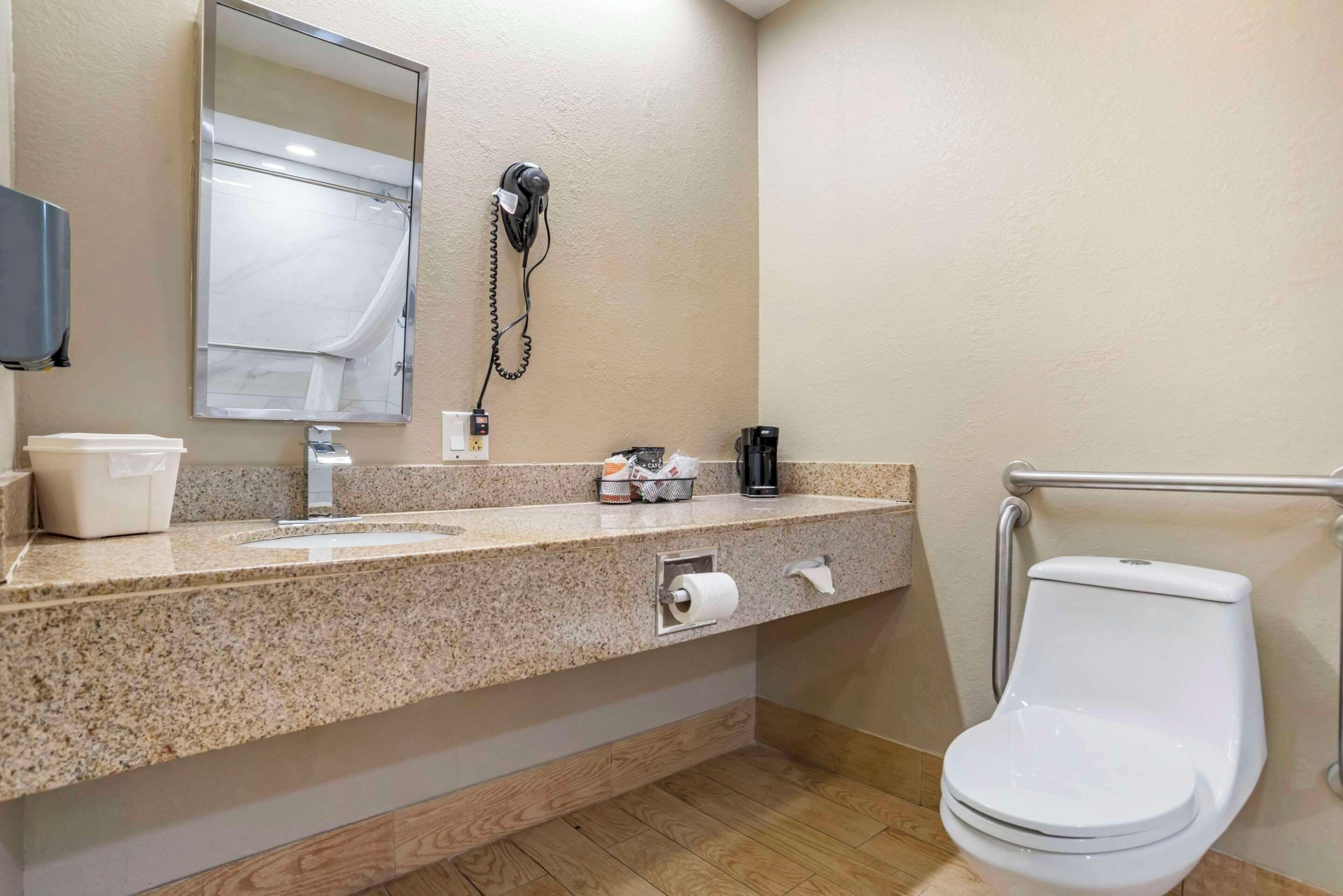 A bathroom with a granite countertop, a sink, a mirror, a toilet, and a hairdryer mounted on the wall.