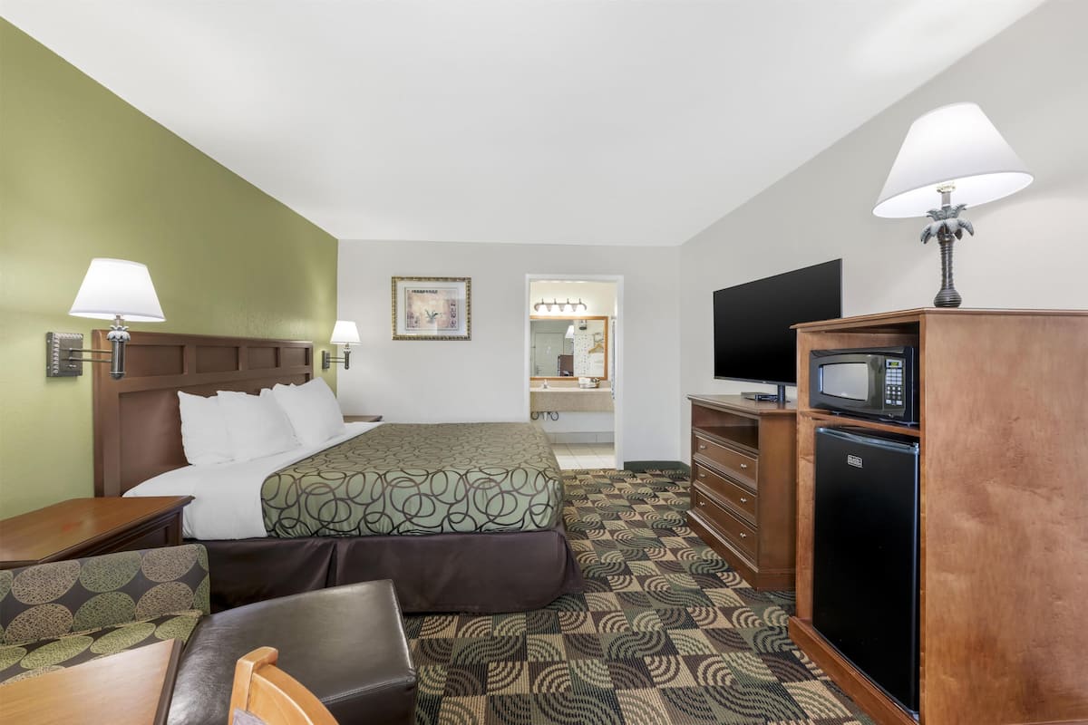A hotel room with a bed, two bedside tables, a TV, a refrigerator, a microwave, a mirror, and a bathroom visible through an open door.