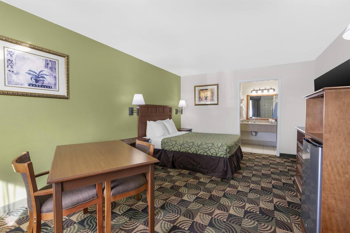 A hotel room with a bed, a desk, a chair, a dresser, a mirror, and a bathroom visible through an open door.