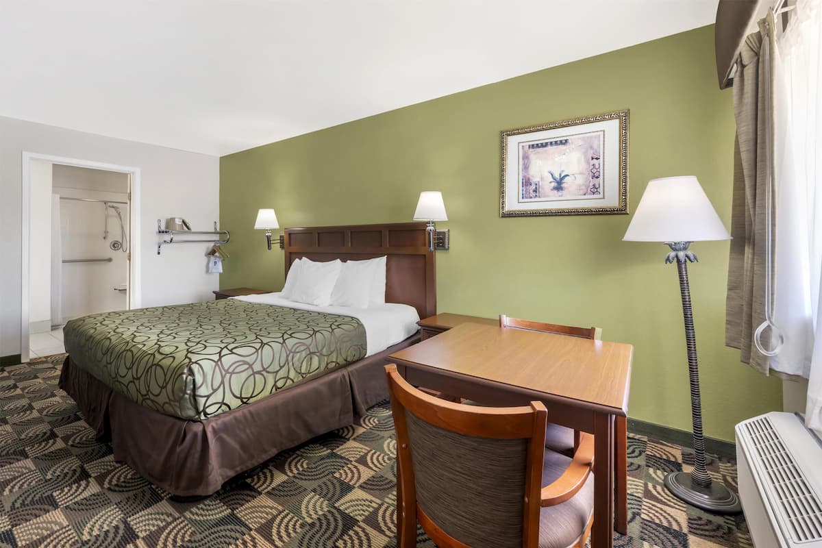 A hotel room with a bed, a wooden table, a chair, a standing heater, and a picture on the wall.