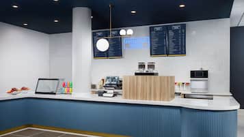 Una moderna zona de recepción con mostrador azul, máquina de café, pantalla digital y cartel de menú.