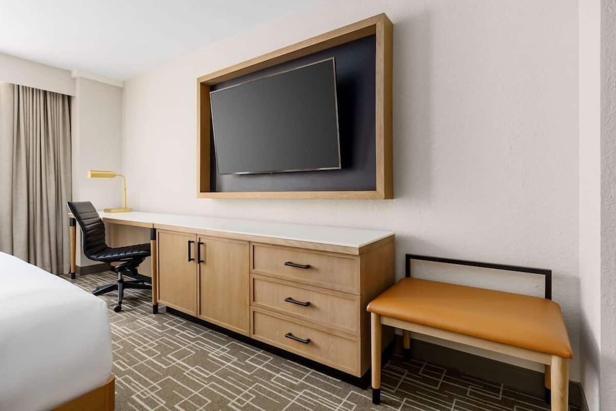 A hotel room with a bed, a desk with a chair, a television mounted on the wall, and a bench.