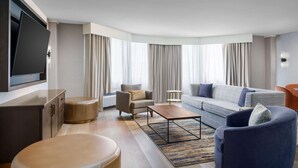 A modern hotel room with a sofa, armchair, ottoman, and a flat-screen TV mounted on the wall.