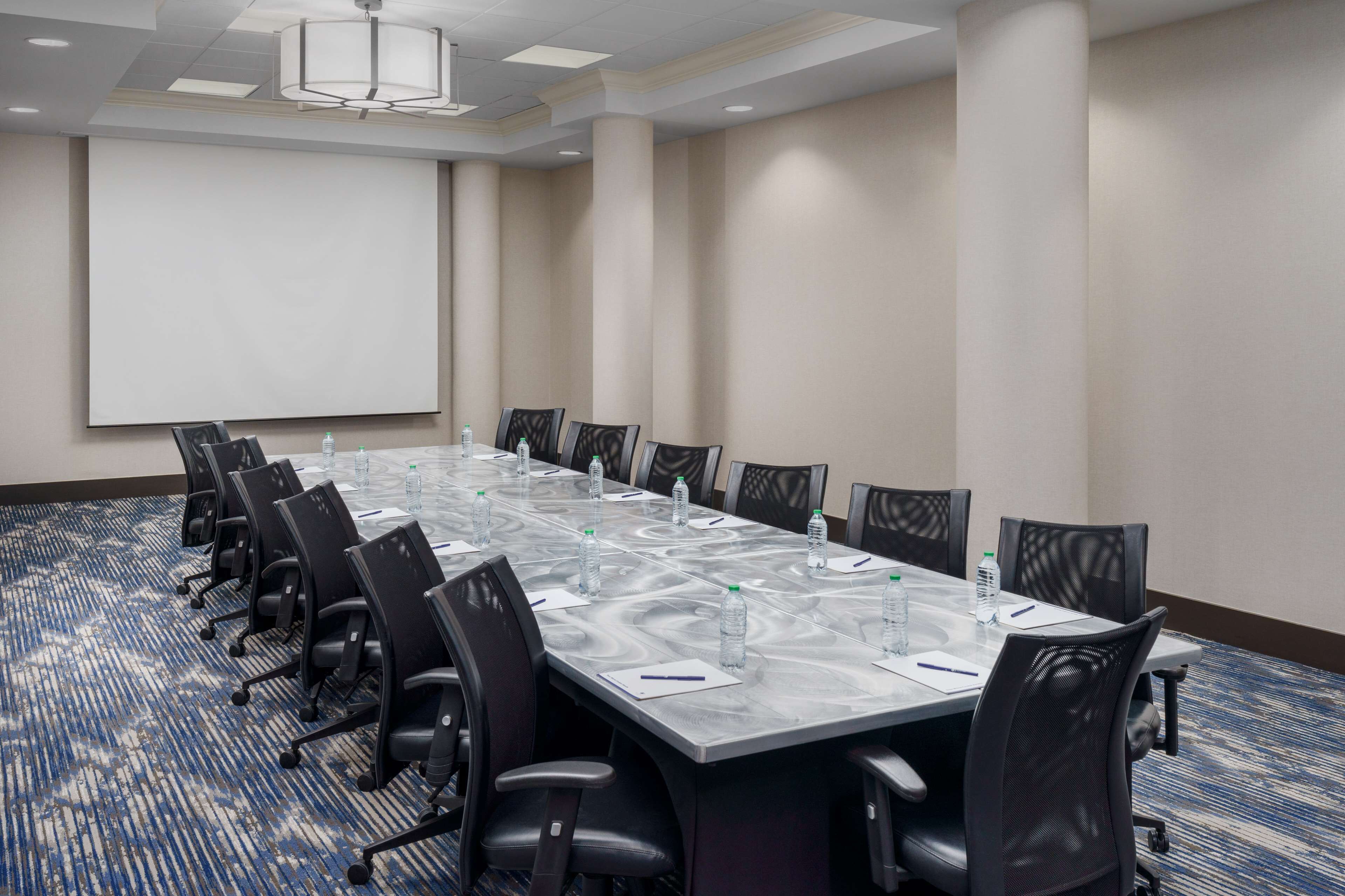 Un salón de conferencias con una larga mesa, sillas, una pantalla de proyector y piso alfombrado.