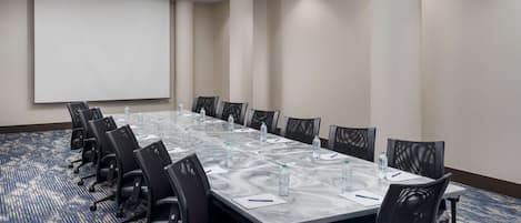 Un salón de conferencias con una larga mesa, sillas, una pantalla de proyector y piso alfombrado.