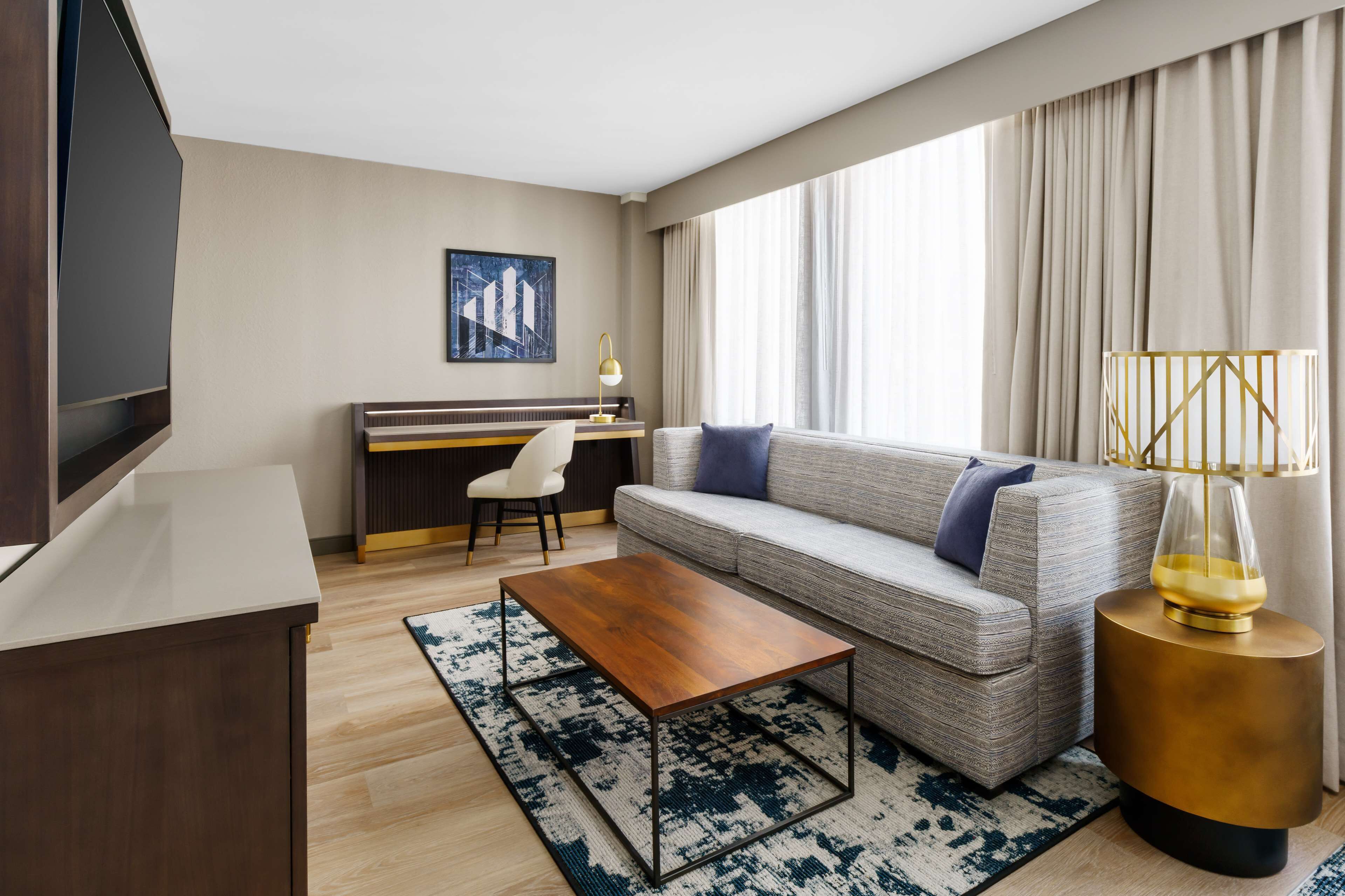 A modern hotel room with a sofa, a wooden coffee table, a desk, and a television.