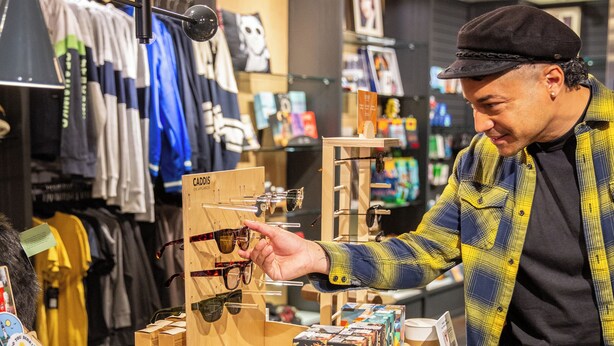 A retail store with clothing racks, a display table with sunglasses, and a person examining sunglasses. - The Edgewater Hotel (Seattle)