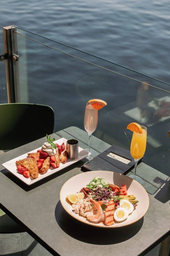 A table with a plate of food and two drinks, a glass of orange juice, and a glass of champagne, with a view of the water in the background.