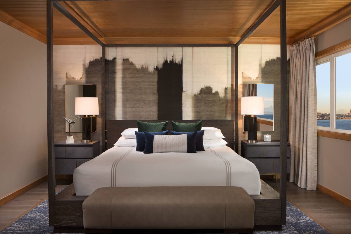 A spacious hotel room with a large bed, two bedside tables with lamps, a view of the sea, and a cityscape painting on the wall.