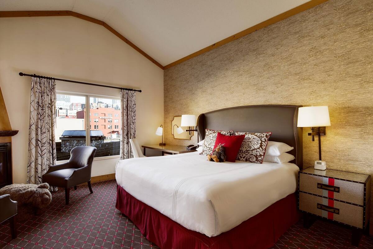 A hotel room with a large bed, a chair, a desk, a lamp, and a view of the city through a window.