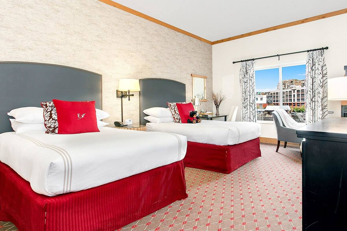 A hotel room with two beds, a desk, a chair, a window with a view of buildings, and a lamp.
