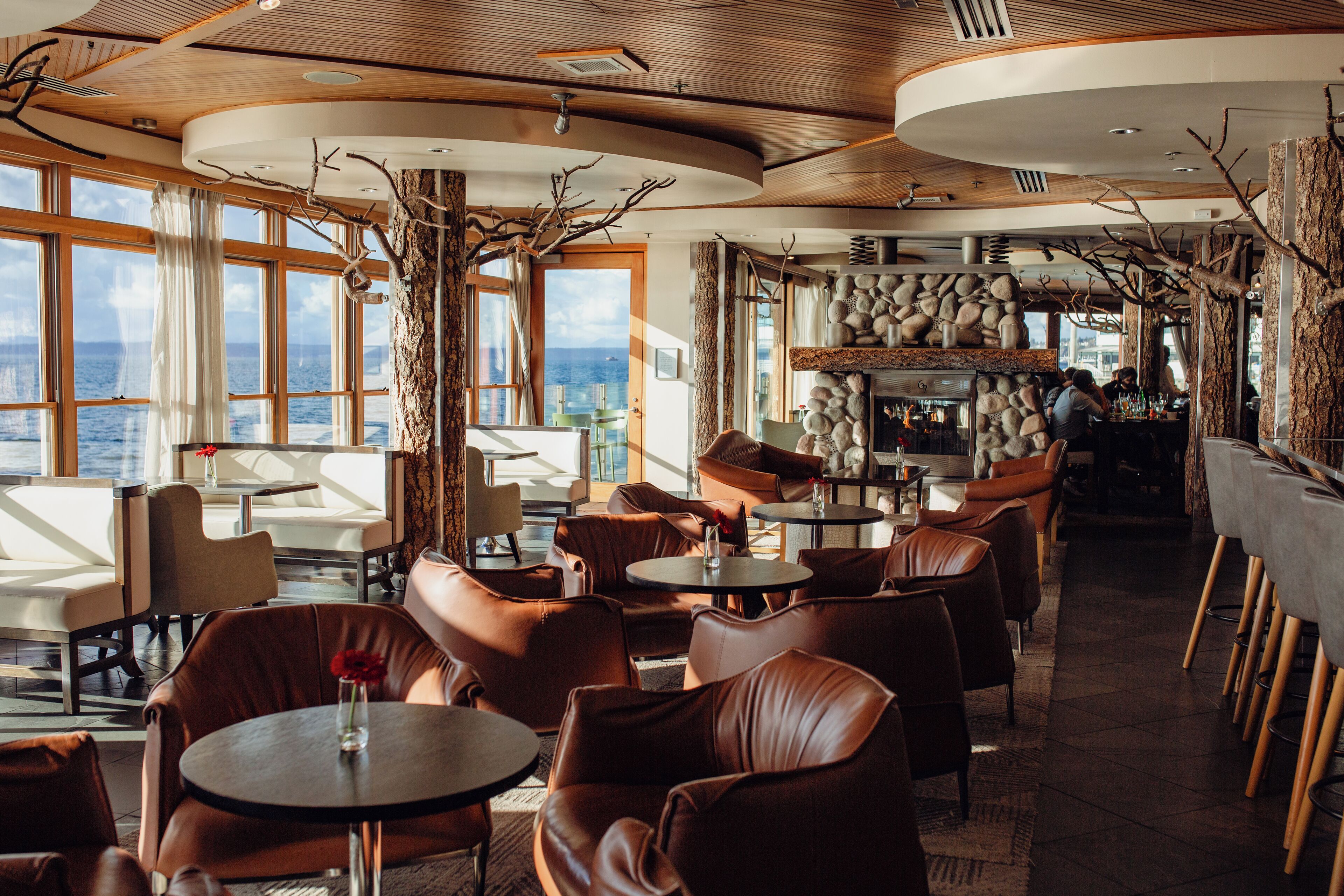 A restaurant with leather seating, a stone fireplace, and large windows overlooking the ocean.