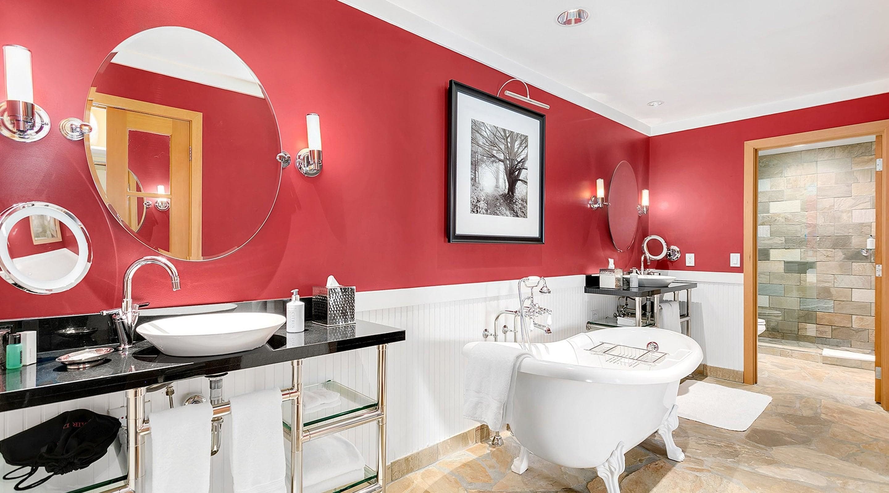 A modern bathroom with a red wall, a white bathtub, a double sink vanity, and a large mirror.