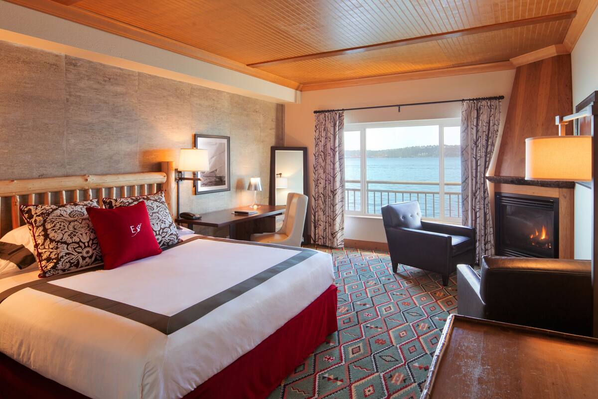 A hotel room with a large bed, a desk, a chair, a fireplace, and a view of the water.