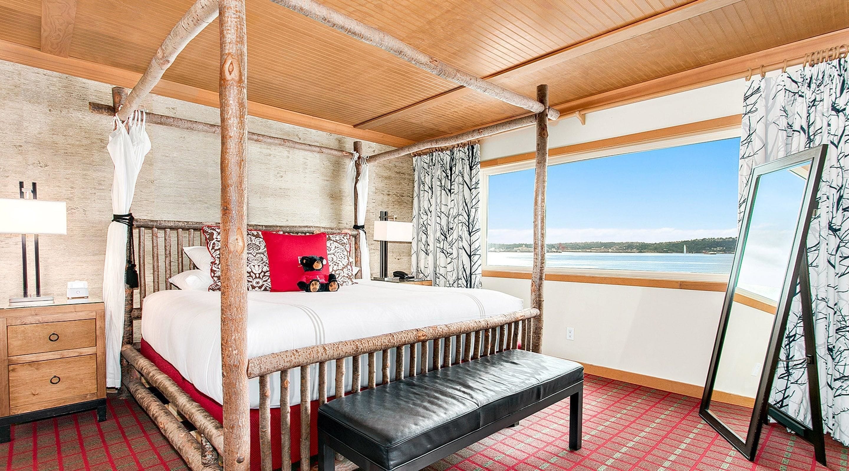 A four-poster bed with a bench, a bedside table, a lamp, and a view of the sea.