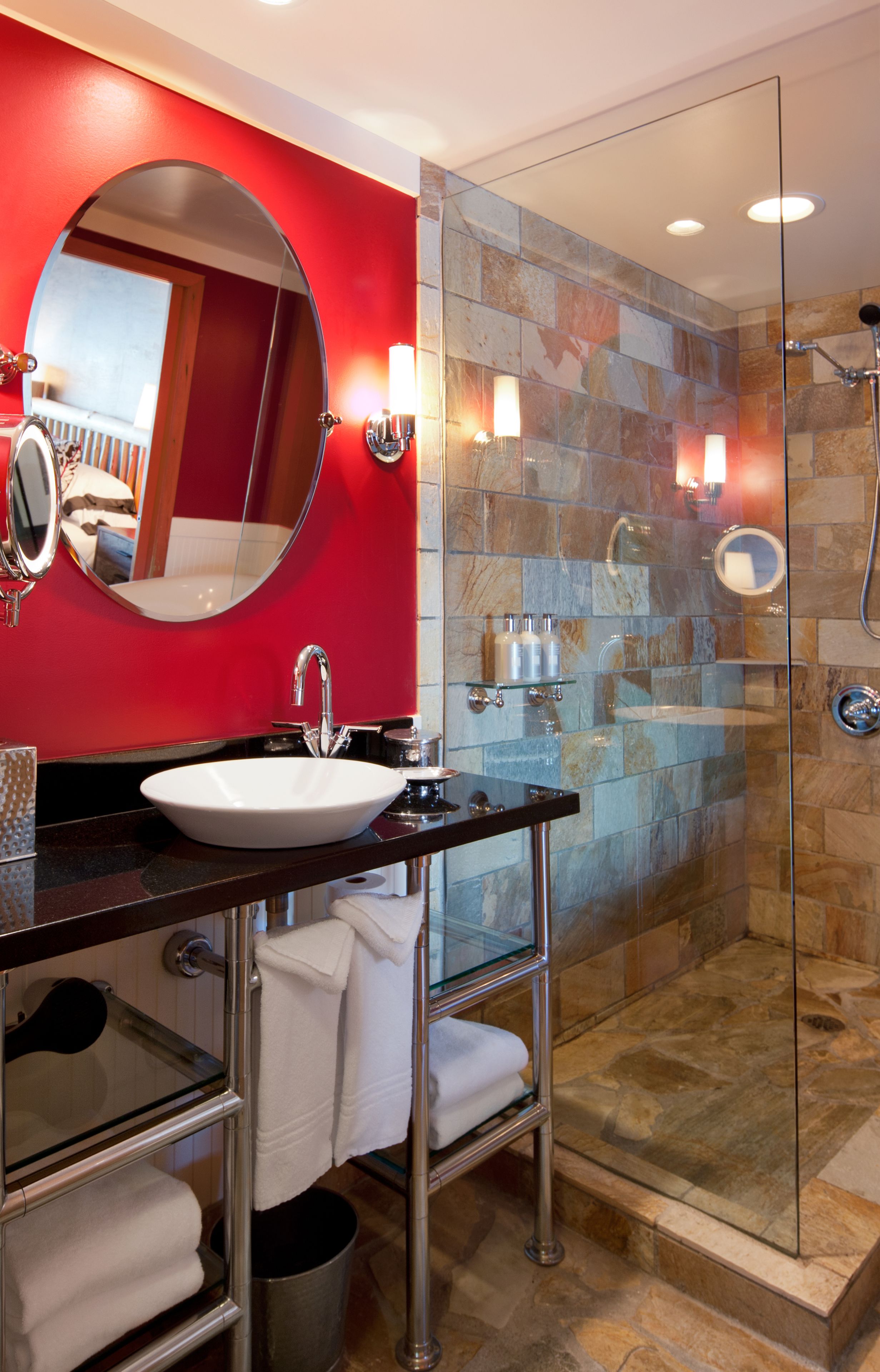 A modern bathroom with a red accent wall, a round mirror, a white sink, and a glass-enclosed shower area.