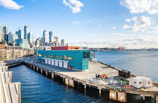 A waterfront dock with a blue building, a city skyline in the background, and a clear sky.