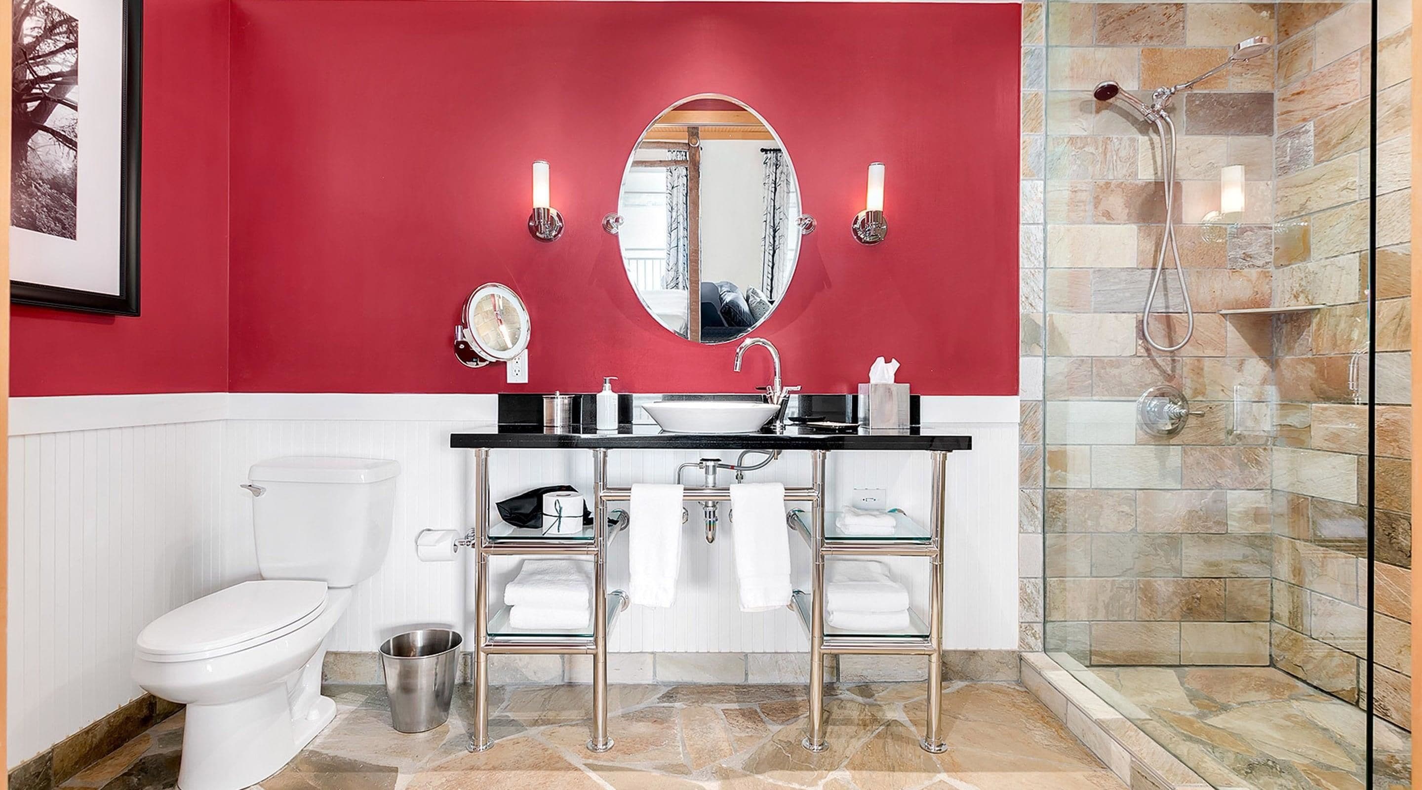 Un baño con pared roja, inodoro blanco, cabina de ducha de vidrio y tocador con espejo y dos luces fijadas a la pared.
