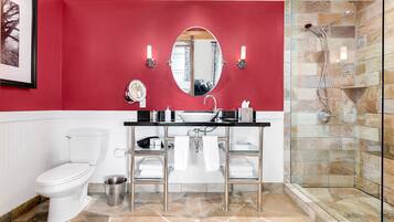 Un baño con pared roja, inodoro blanco, cabina de ducha de vidrio y tocador con espejo y dos luces fijadas a la pared.