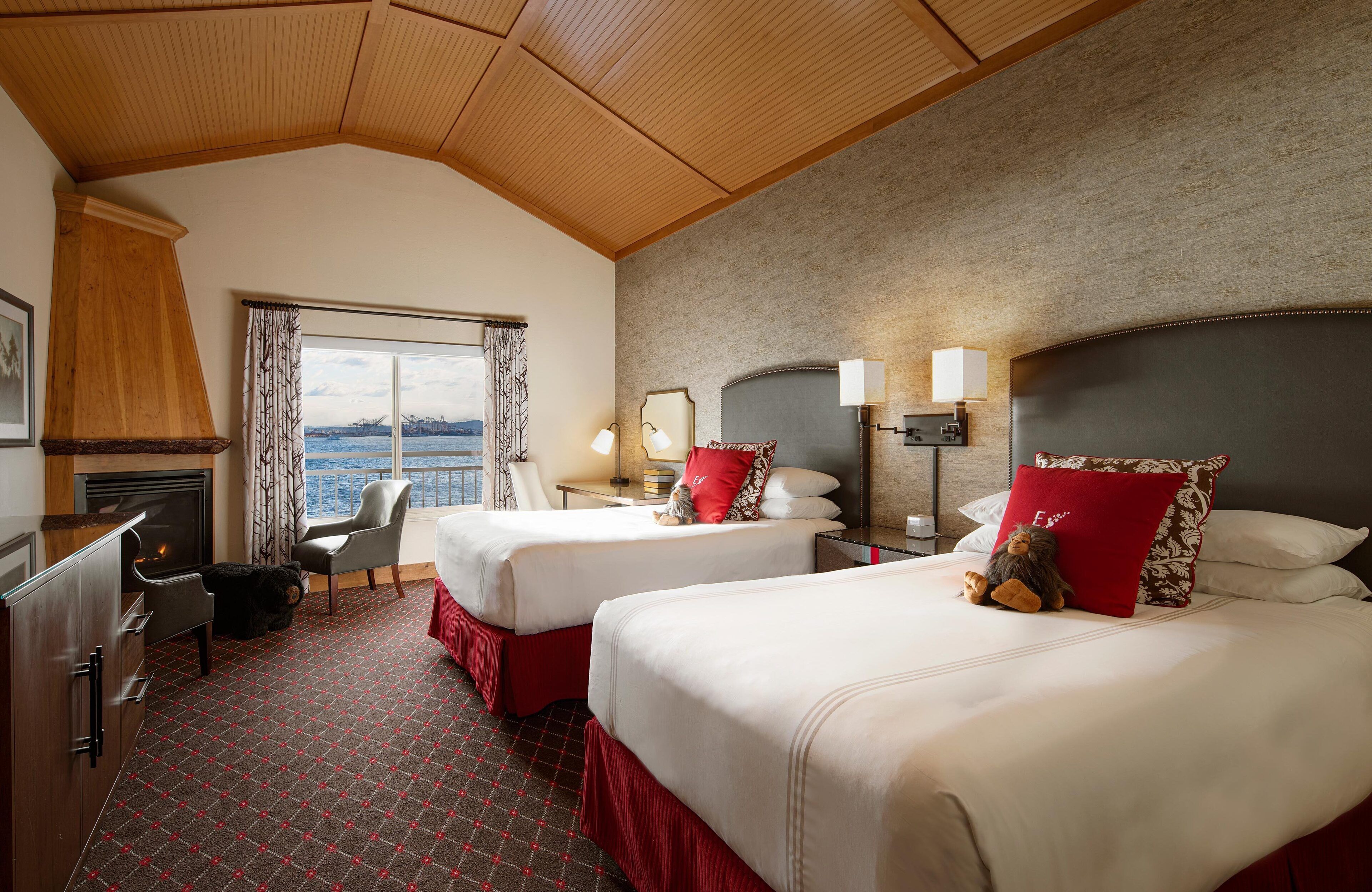 A hotel room with two beds, a desk, a chair, a fireplace, and a view of the water.