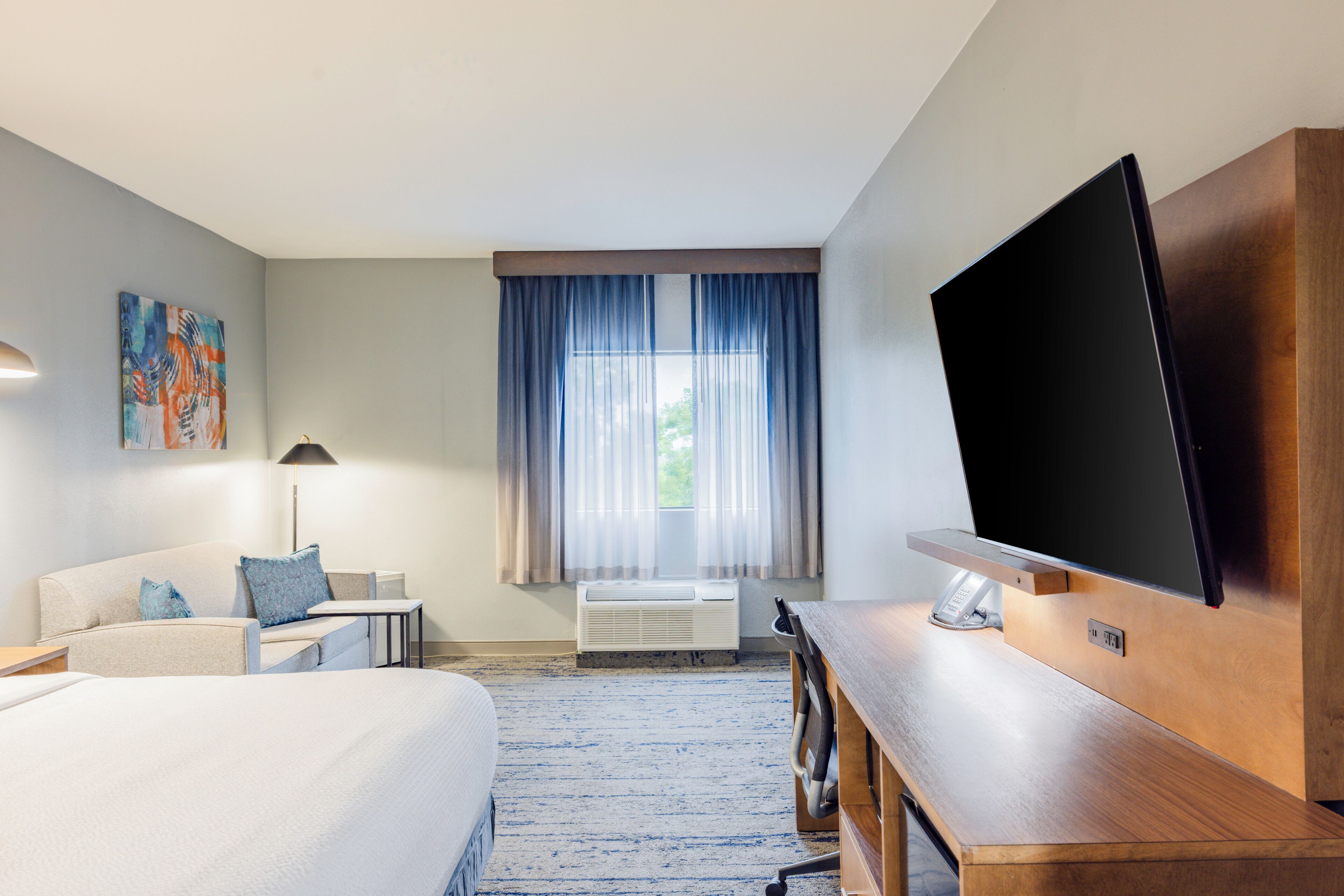 Ein Hotelzimmer mit Bett, Fernseher, Schreibtisch, Stuhl, Lampe, einem Bild an der Wand und einem fenster mit Vorhängen.