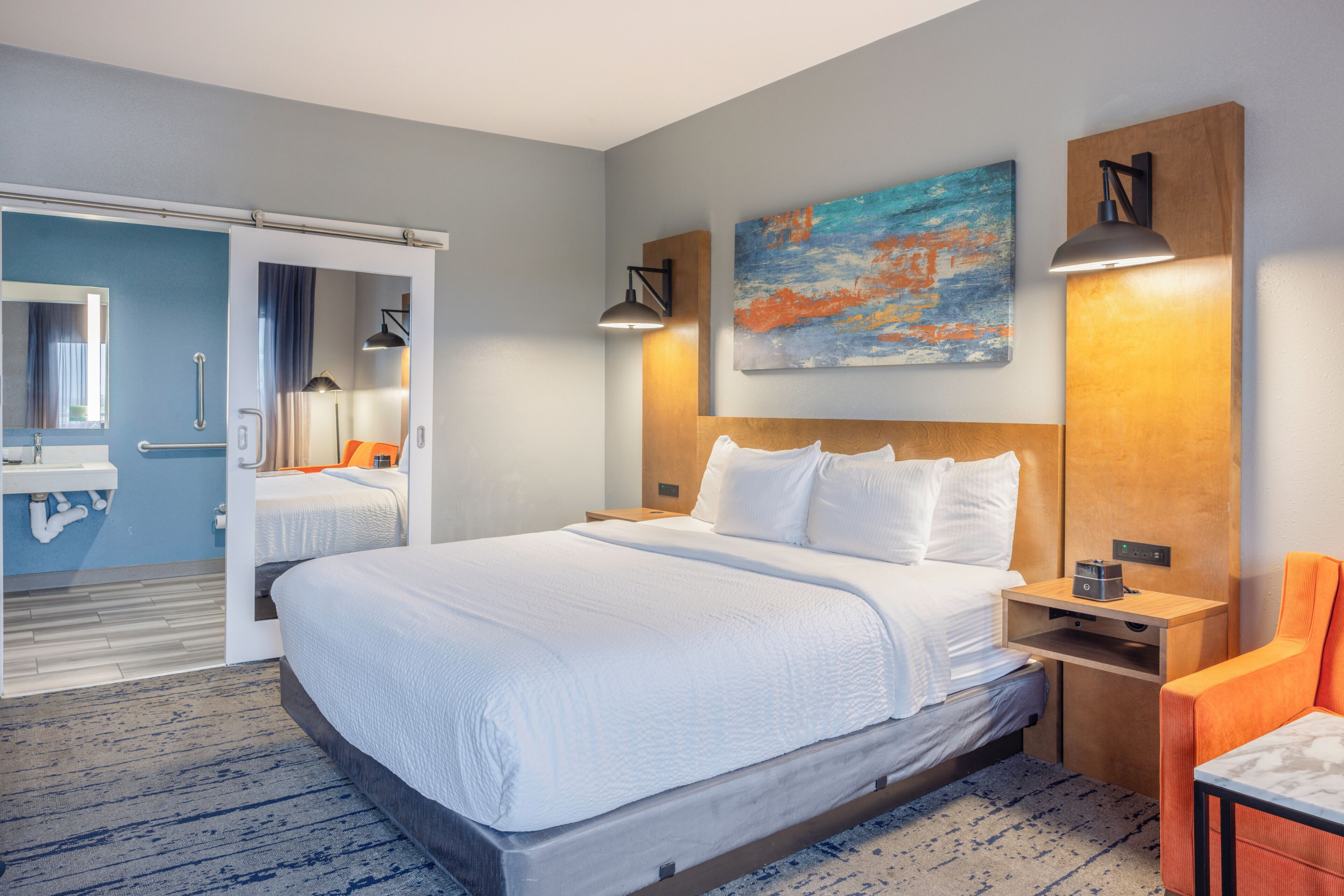 A hotel room with a large bed, a nightstand, a desk, a chair, a painting, and a bathroom with a mirror and sink.