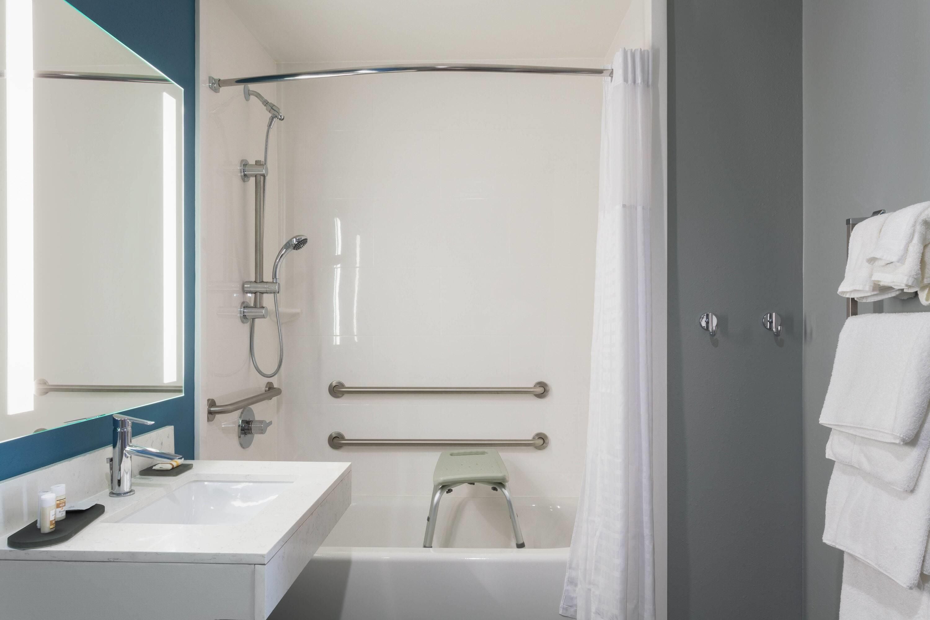 Ein Badezimmer mit weißem Waschbecken, einem Spiegel, einer Dusche mit Duschbrausehandstück, einer Haltestange und einem Handtuchhalter.