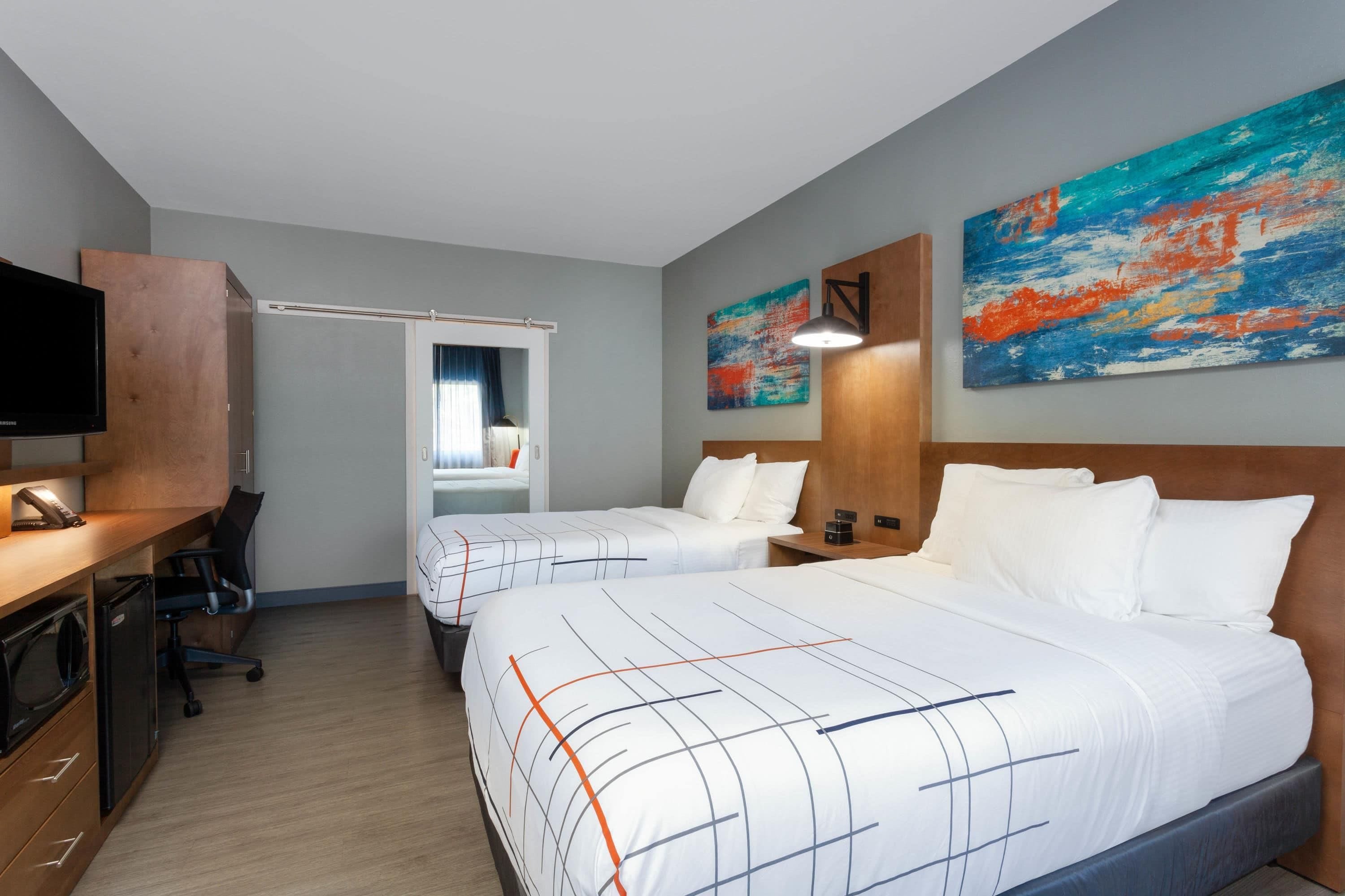 Quarto de hotel com duas camas, uma escrivaninha, televisão e obras de arte nas paredes.