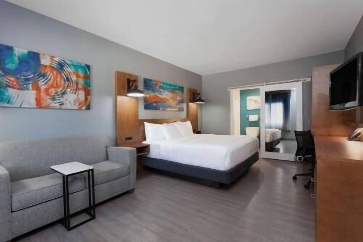 Habitación de hotel con cama, sofá gris, una mesita, obras de arte en las paredes y televisor.