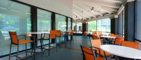 A spacious indoor dining area with high ceilings, large windows, and modern orange chairs and white tables.