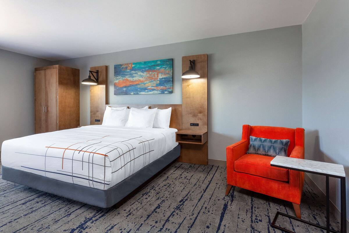 Una habitación de hotel con una cama grande, un sillón rojo, una mesita y un cuadro en la pared.