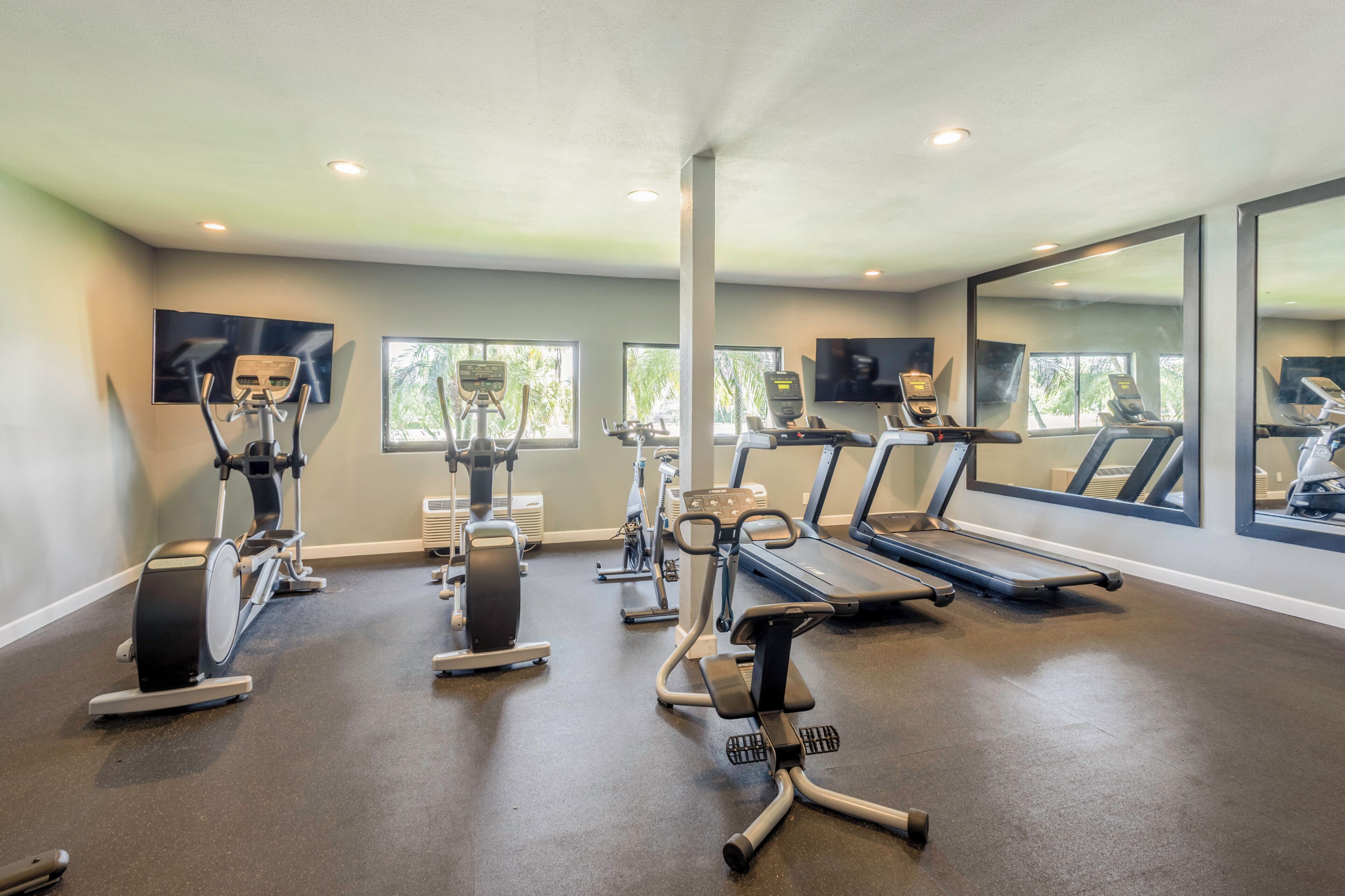 A gym with various exercise equipment, large windows, and a flat-screen TV mounted on the wall.