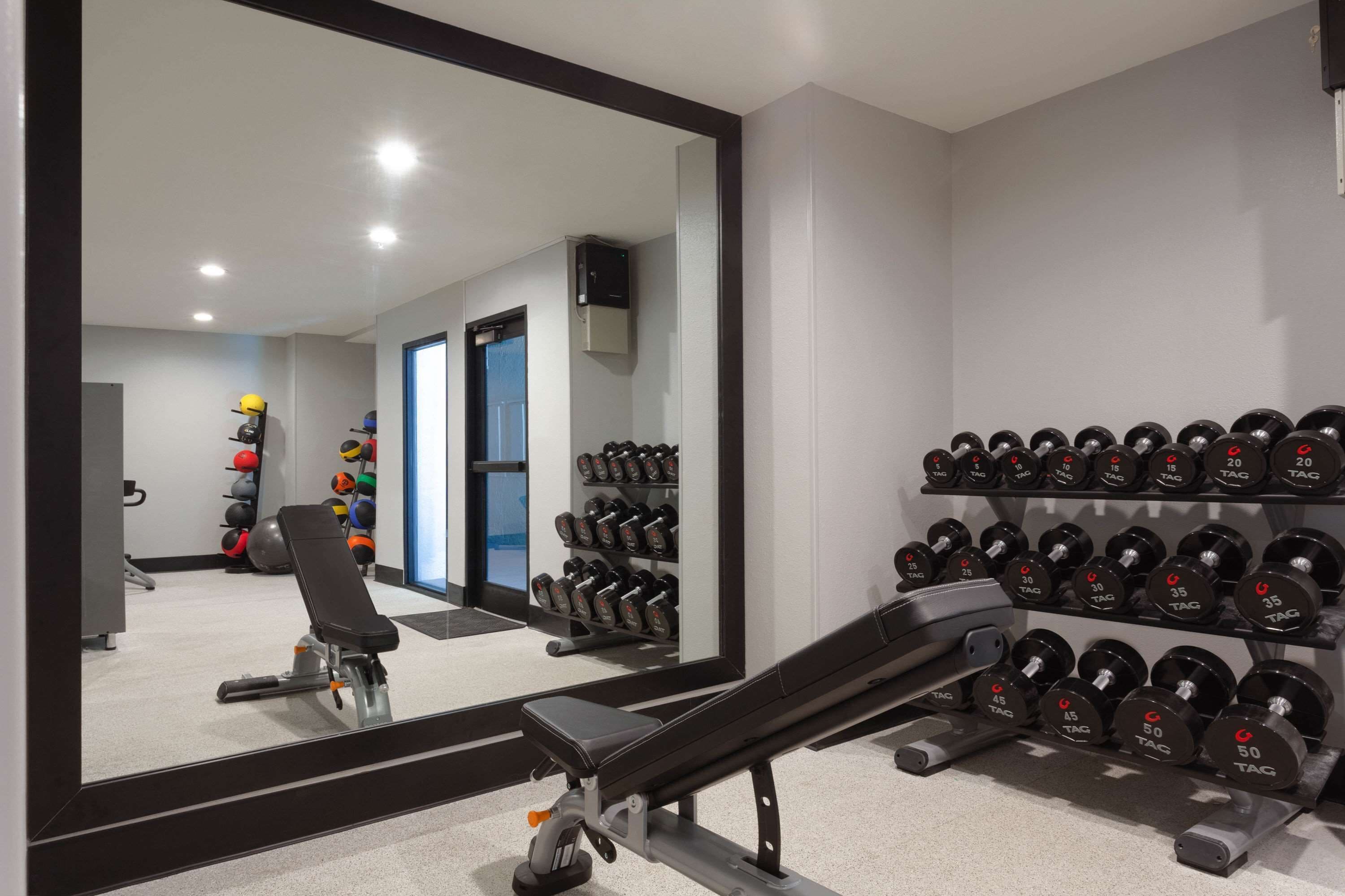 Un gimnasio con una prensa de banca, mancuernas y un espejo que refleja el espacio.