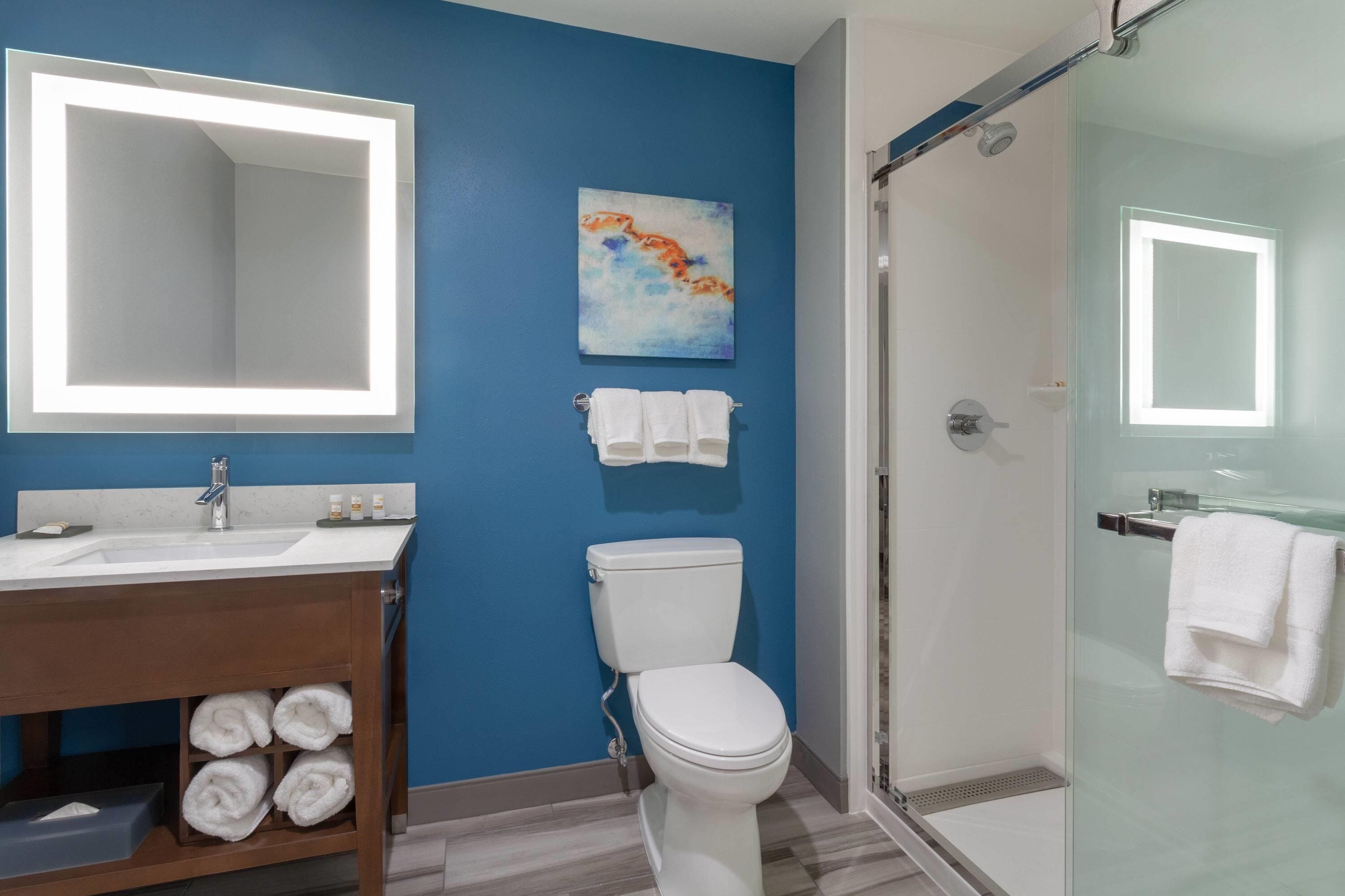 Un baño con pared azul, inodoro blanco, cabina de ducha con mampara de vidrio y tocador de madera con toallas.