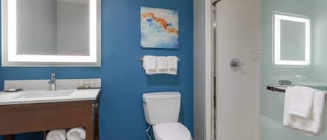 Un baño con pared azul, inodoro blanco, cabina de ducha con mampara de vidrio y tocador de madera con toallas.