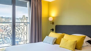 A hotel room with a large bed, yellow and grey pillows, a grey headboard, a window with a curtain, and a view of buildings outside. - Best Western Plus Hôtel de Dieppe 1880 (Rouen)