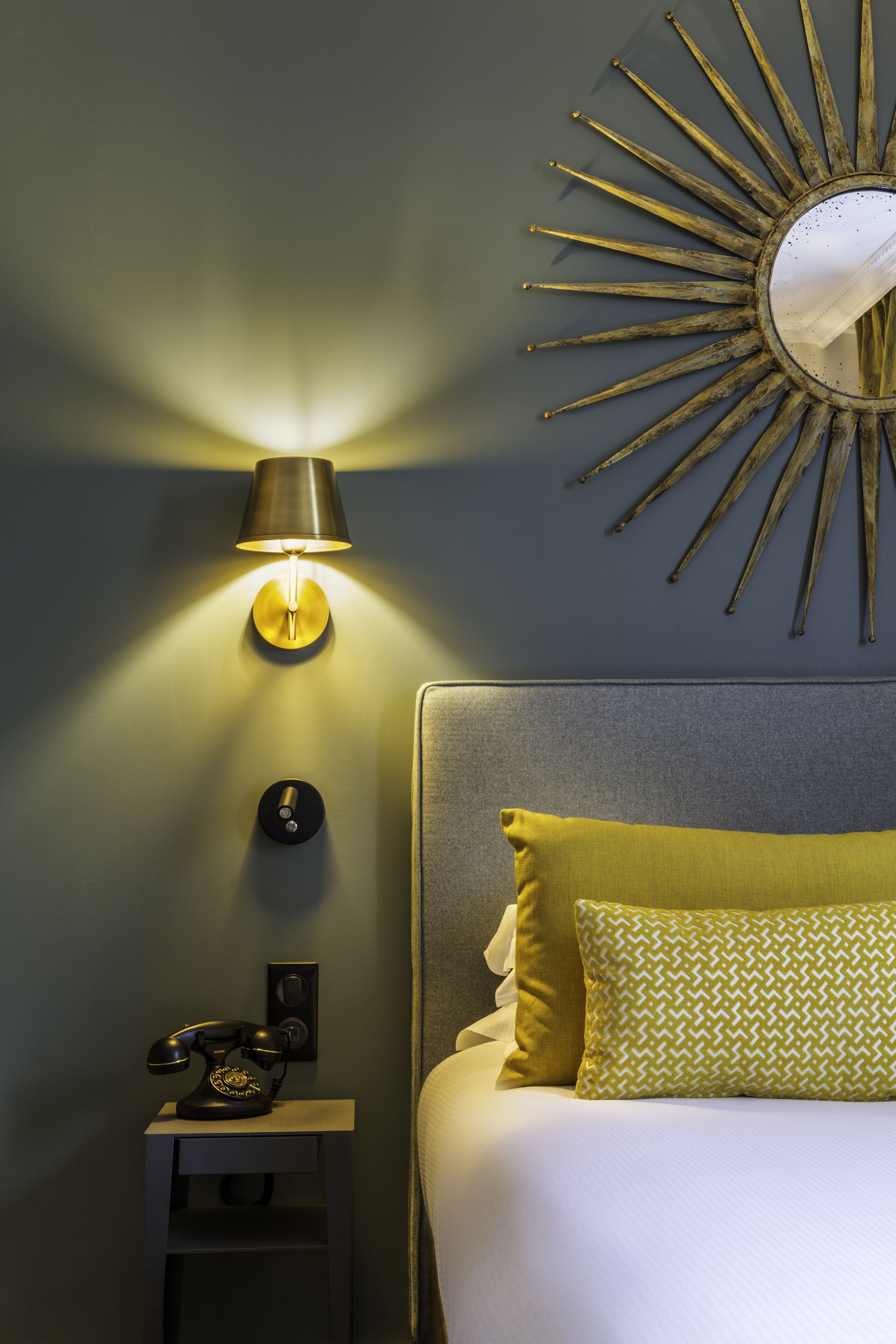 Un lit bien fait, agrémenté d’un coussin à motifs, une lampe fixée au mur, un téléphone d’époque et un miroir en forme de soleil accroché au mur.