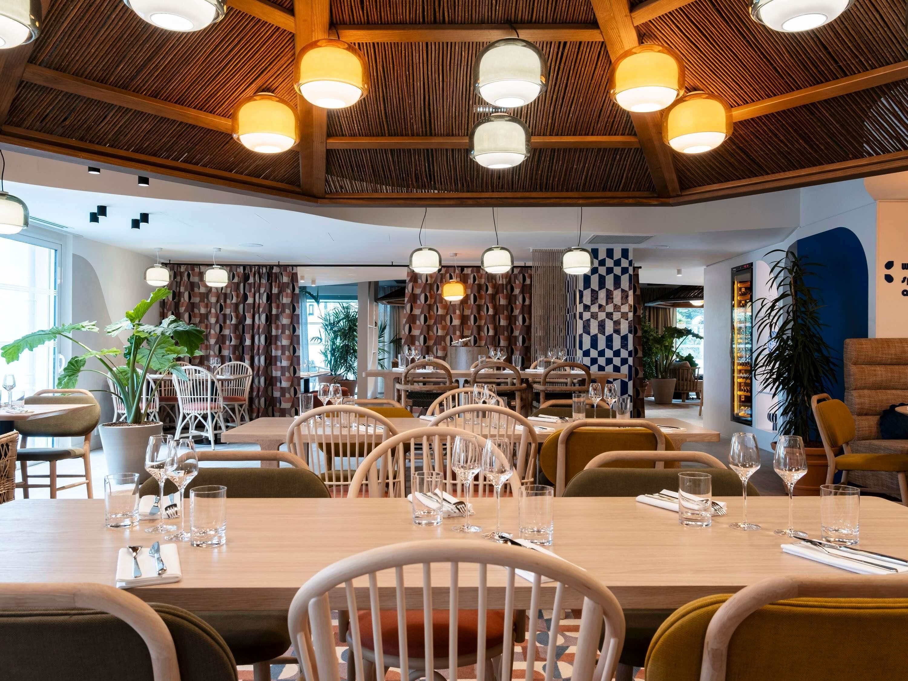 En restaurang med synliga träbjälkar i taket, hängande lampor och ett rutmönstrat väggmåleri. Borden är dukade med stolar och vinglas.