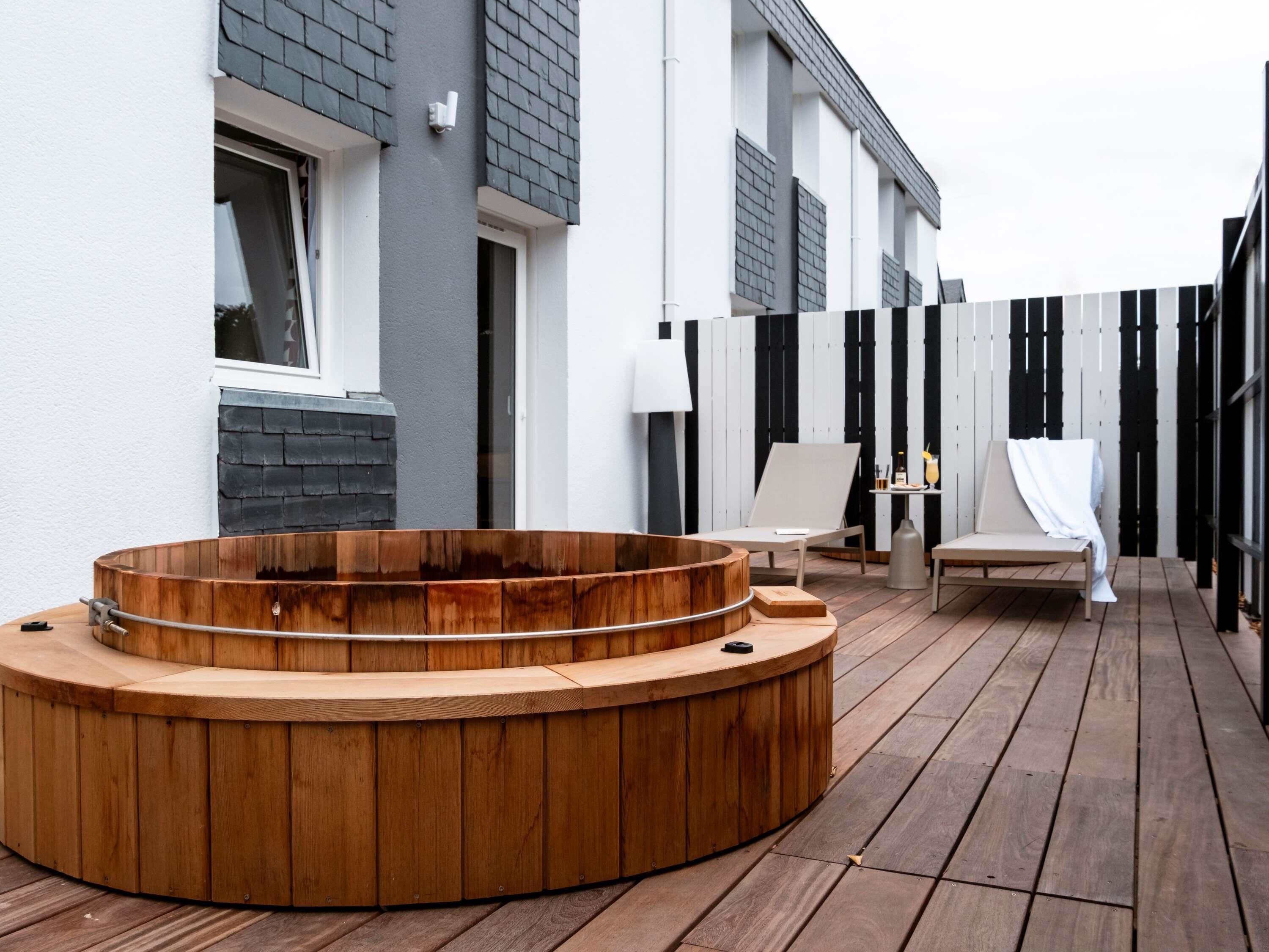 Un jacuzzi de madera en una terraza de madera, un edificio blanco con contraventanas grises y una valla negra y blanca.