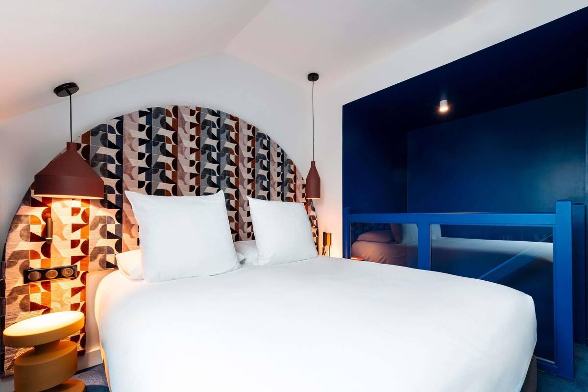 A modern hotel room with a large bed, geometric-patterned headboard, and a blue accent wall.